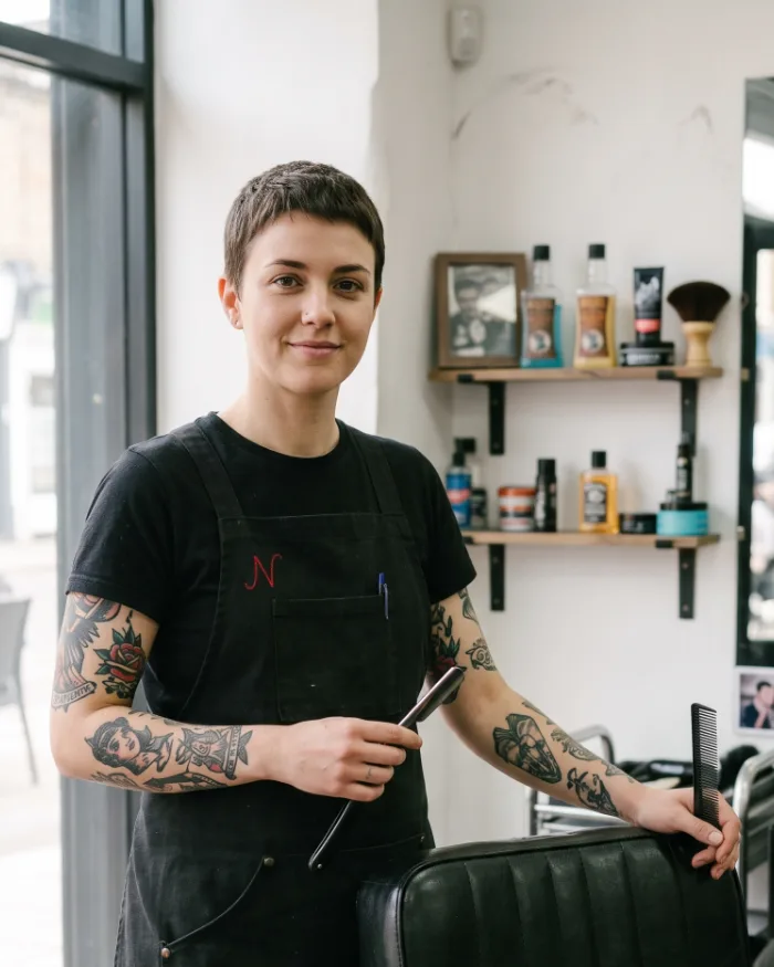 Dee, a barber with short cropped hair, beside her chair